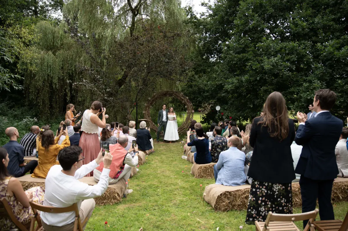 An outdoor ceremony under a flower arch, guests seated on hay bales among the trees