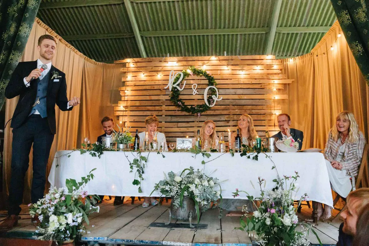 A speech beside the top table in the barn, under festoon lights and a foliage wreath