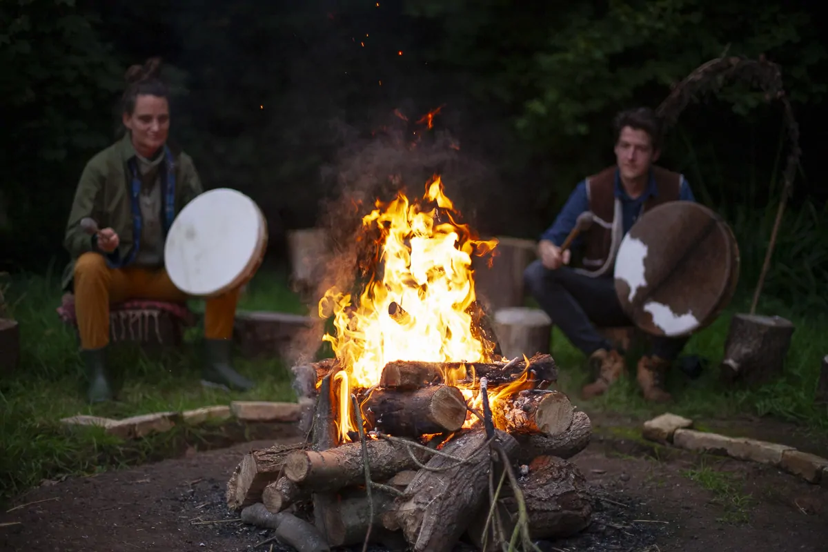 Two drummers playing beside a bonfire at dusk